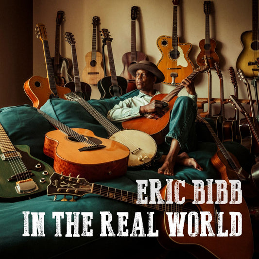Eric Bibb poses with guitars, album title “In The Real World” visible.