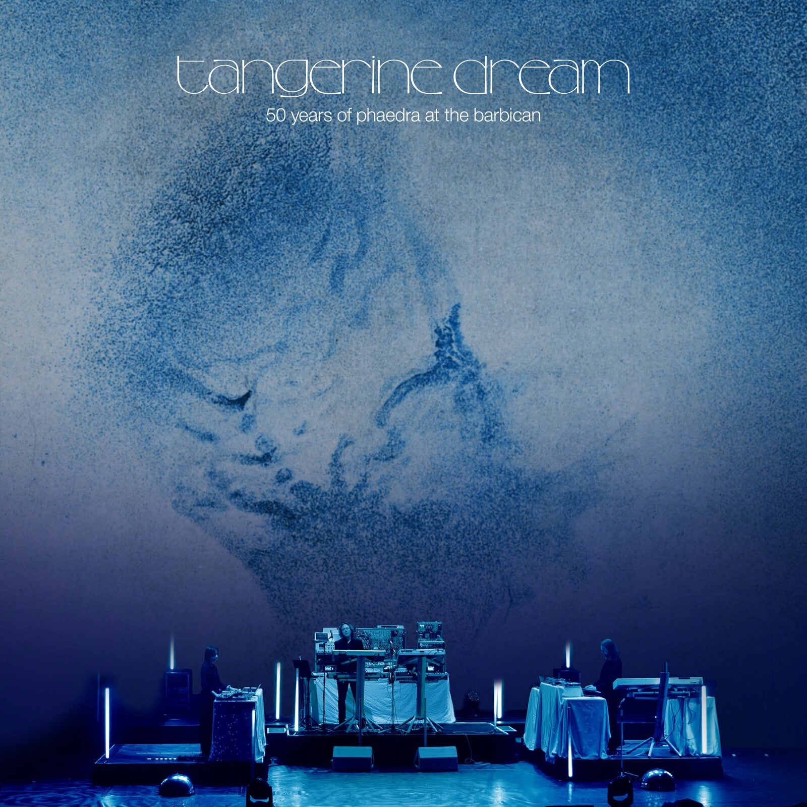 Tangerine Dream performing on stage with a large blue abstract projection in the background.