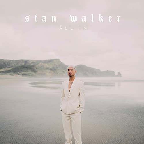 Stan Walker in a light suit standing on a beach with mountains in background.