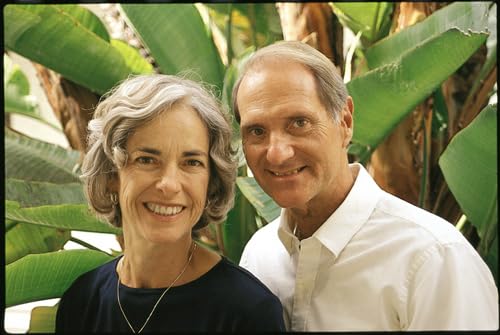 Two smiling adults stand close together with lush green foliage behind them.