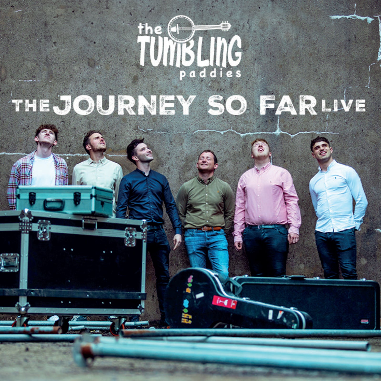 Six men stand in front of a concrete wall with musical equipment cases nearby.