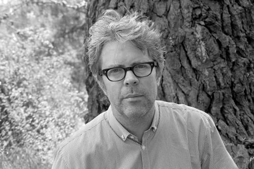 Black and white portrait of a man with glasses, leaning against a tree.