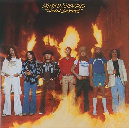 Seven men stand in front of a wall of flames wearing casual clothing.
