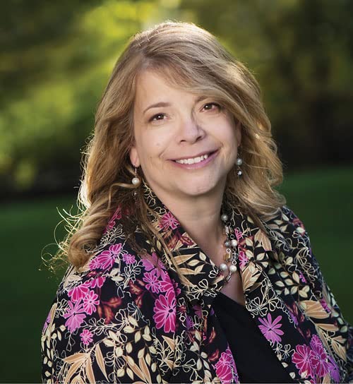 Woman with blonde hair smiling, wearing a floral patterned jacket.