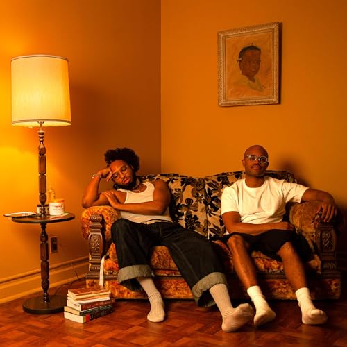 Two men sit on a patterned sofa in a room with orange walls.