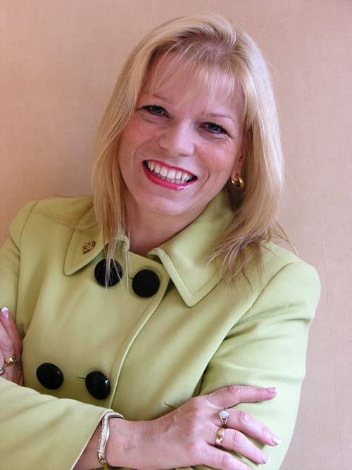 Blonde woman smiling, wearing a green jacket and jewelry.