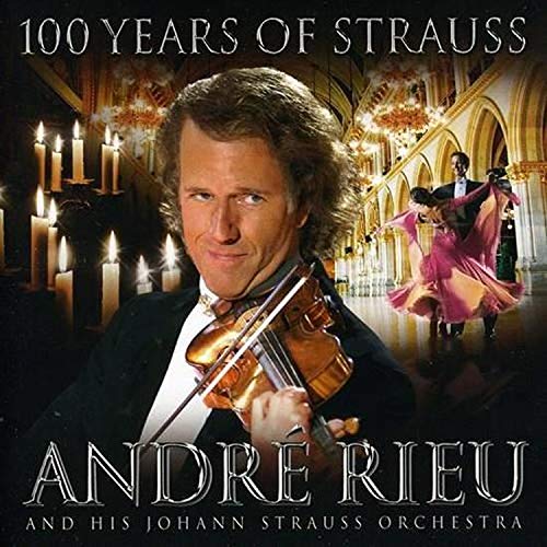 Andre Rieu holding a violin, with dancers and a grand staircase backdrop.