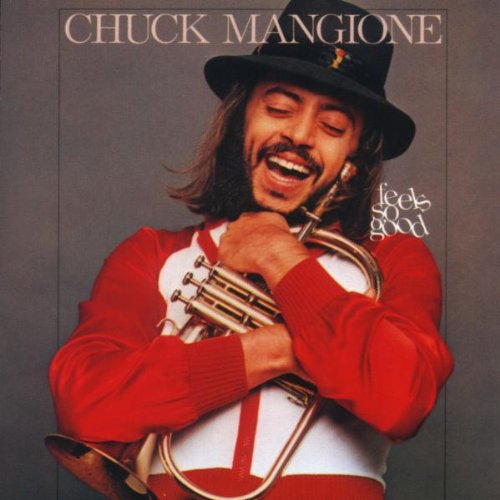 Chuck Mangione smiling, holding a flugelhorn, wearing a hat and red sweater.