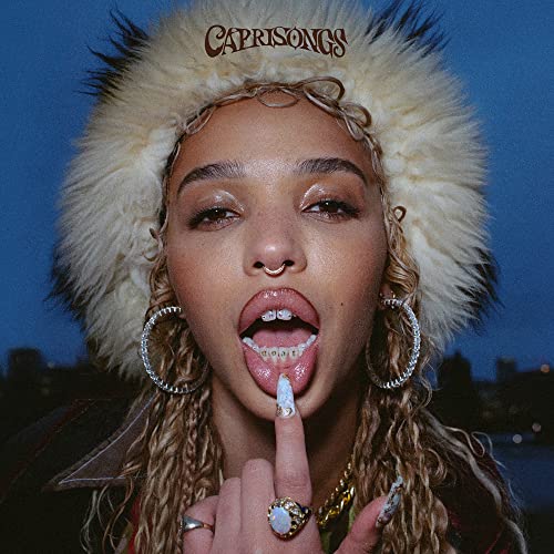 FKA Twigs wearing a fur hat with her finger touching her tongue and teeth.