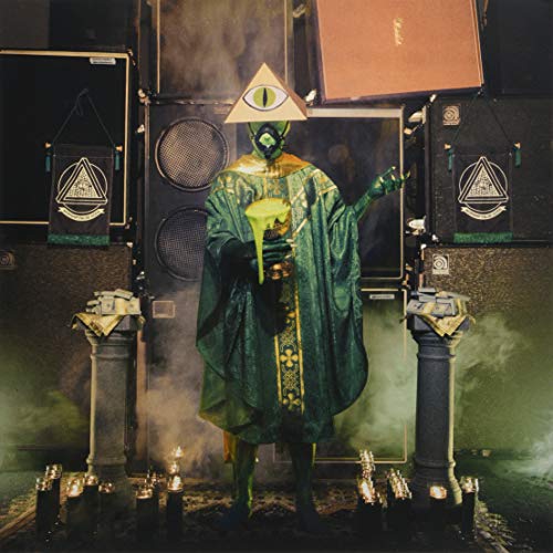 Green reptilian figure with a single eye, standing before speakers.