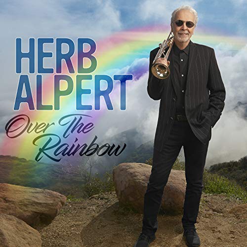 Herb Alpert holding a trumpet, with a rainbow and cloudy sky backdrop.