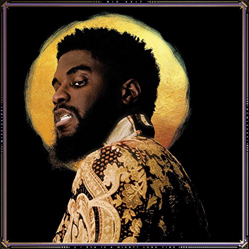Man with short hair and beard wearing ornate gold shirt against black background.