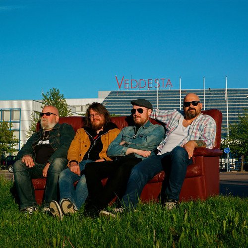 Four men posing on a red couch in front of a building.