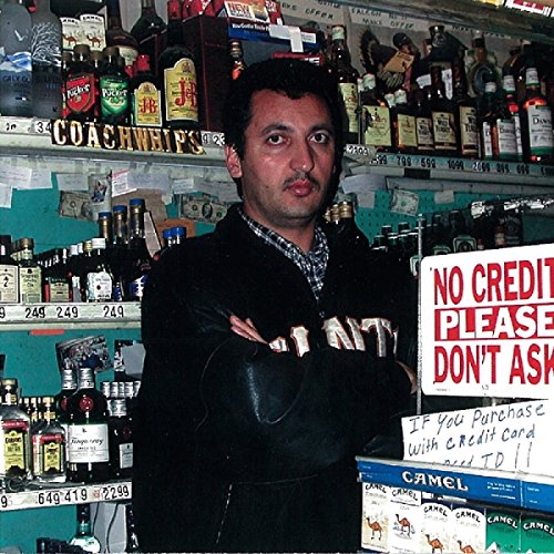 Man with a mustache stands in front of a bar’s liquor shelves.
