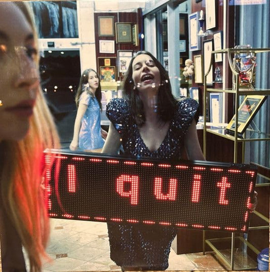 Three women with a glowing red sign reading “I quit” indoors.