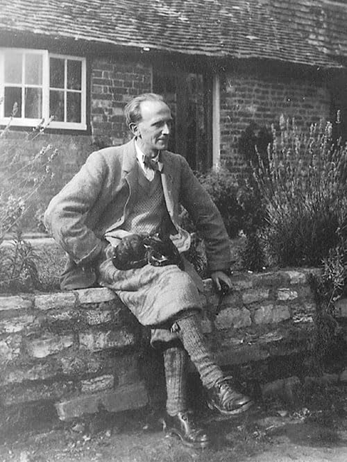 A.A. Milne sits on a stone wall in front of a brick house.