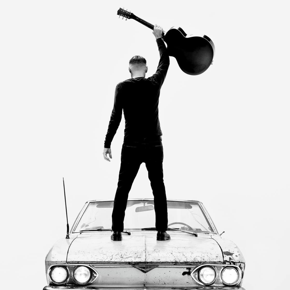 Black and white image of a man with a guitar on a car roof.