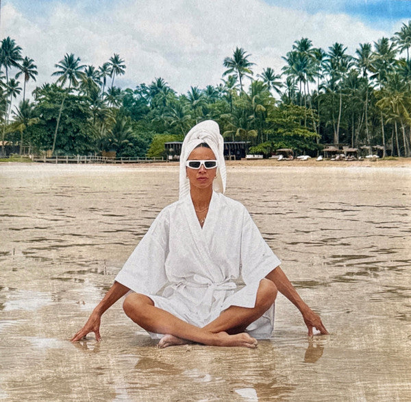 Woman in white robe and sunglasses sits in shallow water.