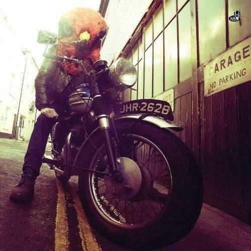 Person in a large furry bird head mask sits on a vintage motorcycle.