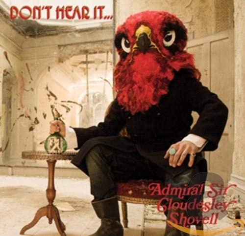 A person in a red bird mask and black suit sitting in a room.