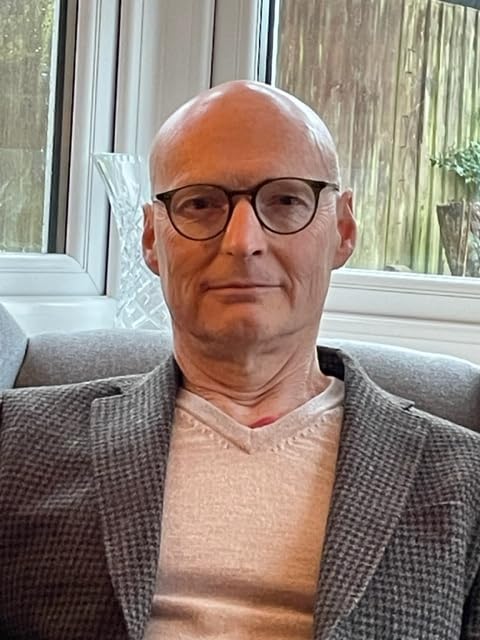 A bald man with glasses wearing a grey blazer and beige v-neck shirt.