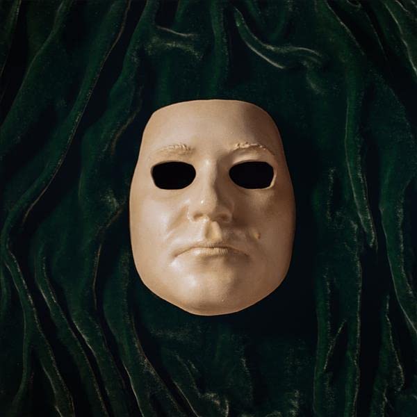 Pale human mask with dark eye sockets against dark green fabric.