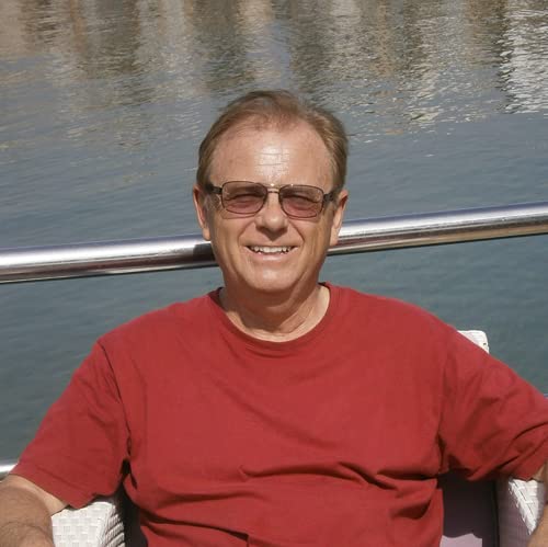A smiling man with glasses wearing a red shirt, sitting in front of water.