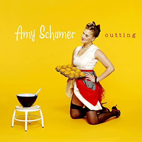 Amy Schumer kneels on a yellow background holding a muffin tin, wearing an apron.