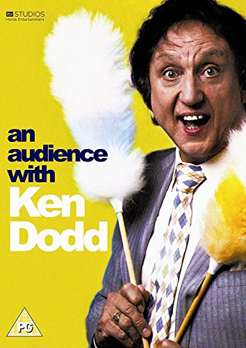 Ken Dodd smiling while holding two feather dusters against a bright yellow background.