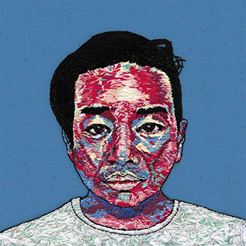 A man with short black hair and a white shirt, embroidered artwork on blue background.