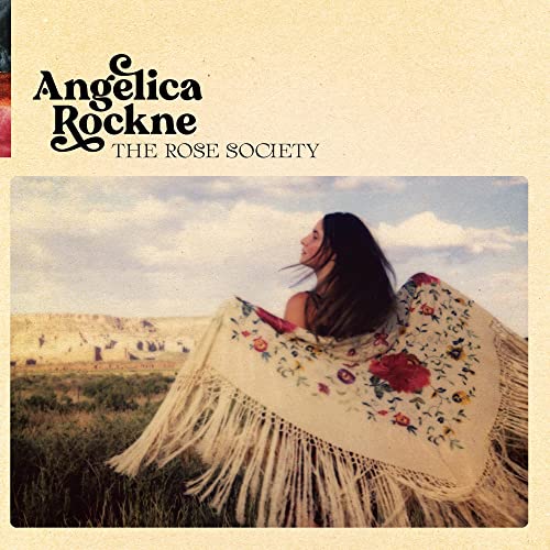 A woman wearing a floral fringed shawl stands in a field under a cloudy sky.