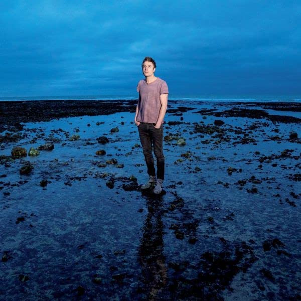 Man standing on a rocky beach under a blue, overcast sky.
