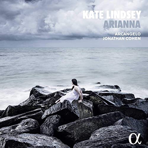 CD cover: Woman in white dress sits on rocky shore under cloudy sky.
