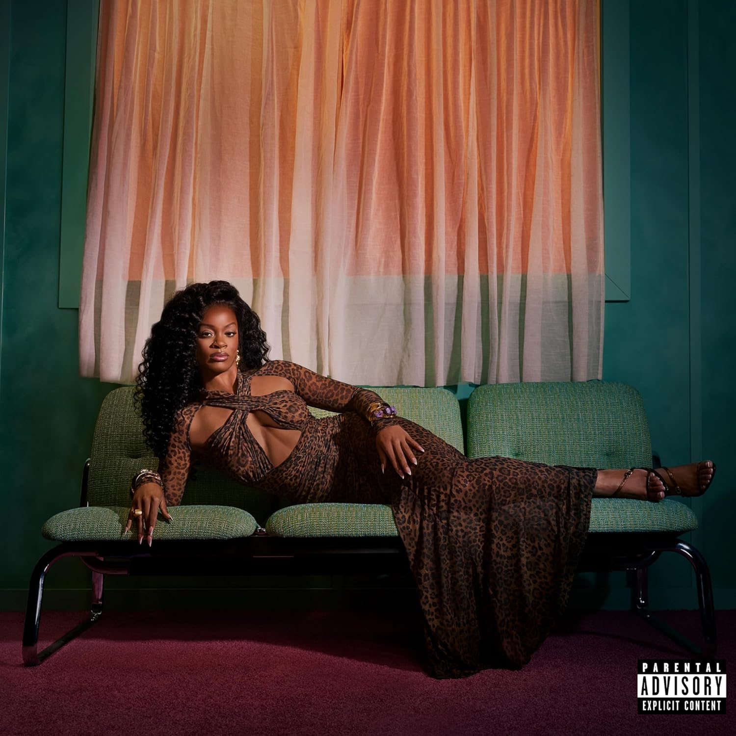 Ari Lennox poses on a green couch with orange curtains behind her.