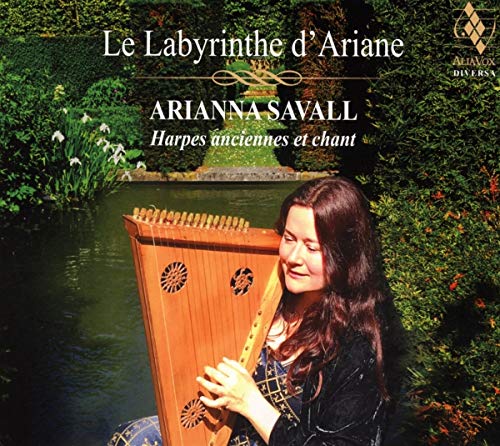 Arianna Savall holding a harp by a pond, surrounded by greenery and flowers.