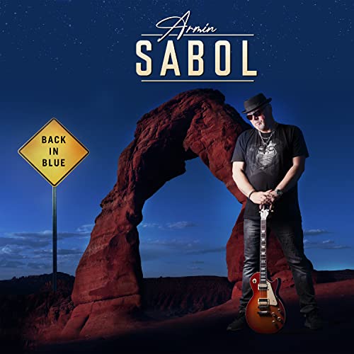 Armin Sabol stands with a guitar before a rock arch and yellow road sign.