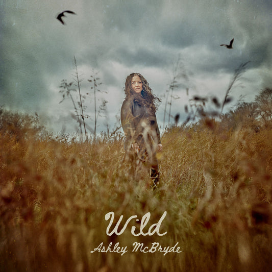 Ashley McBryde stands in a field of tall grass under a cloudy, overcast sky.