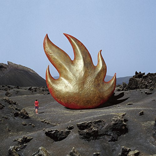A person stands before a large, golden, flame-shaped sculpture in a volcanic landscape.