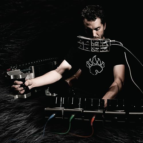 A man wearing a bear-print shirt performs using custom-built industrial mechanical musical instruments.