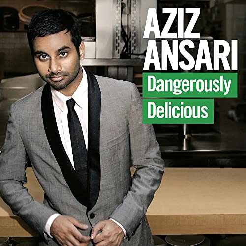 Aziz Ansari wearing a grey suit and black tie in front of a kitchen counter.