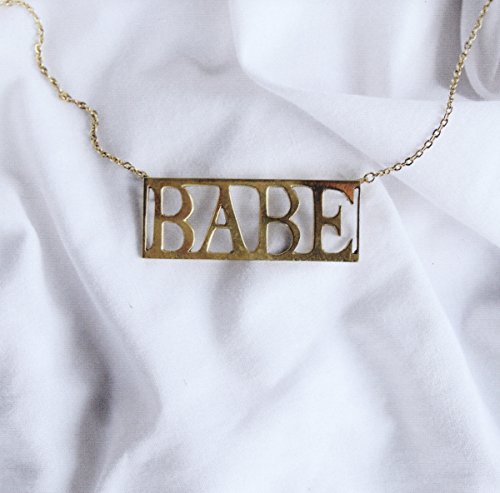 Gold necklace with "BABE" pendant on white fabric, centered and prominent.