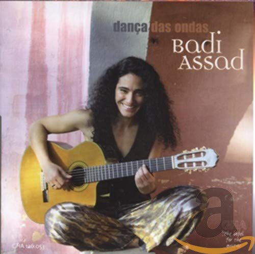 Badi Assad sitting cross-legged, playing an acoustic guitar.
