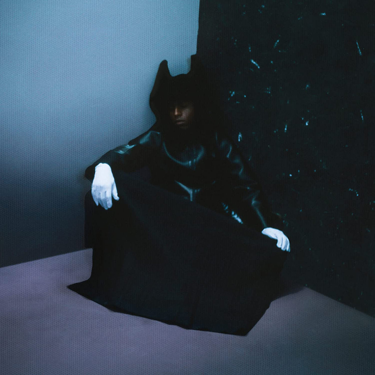 Figure in dark clothing and gloves sits in a shadowed room.