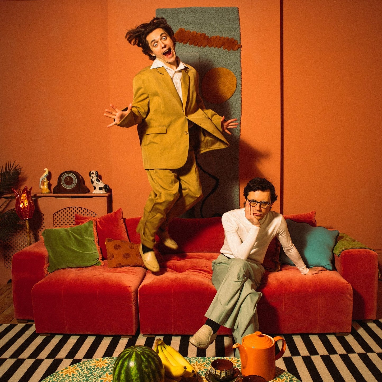Two men in suits pose on a red couch, with fruit nearby.