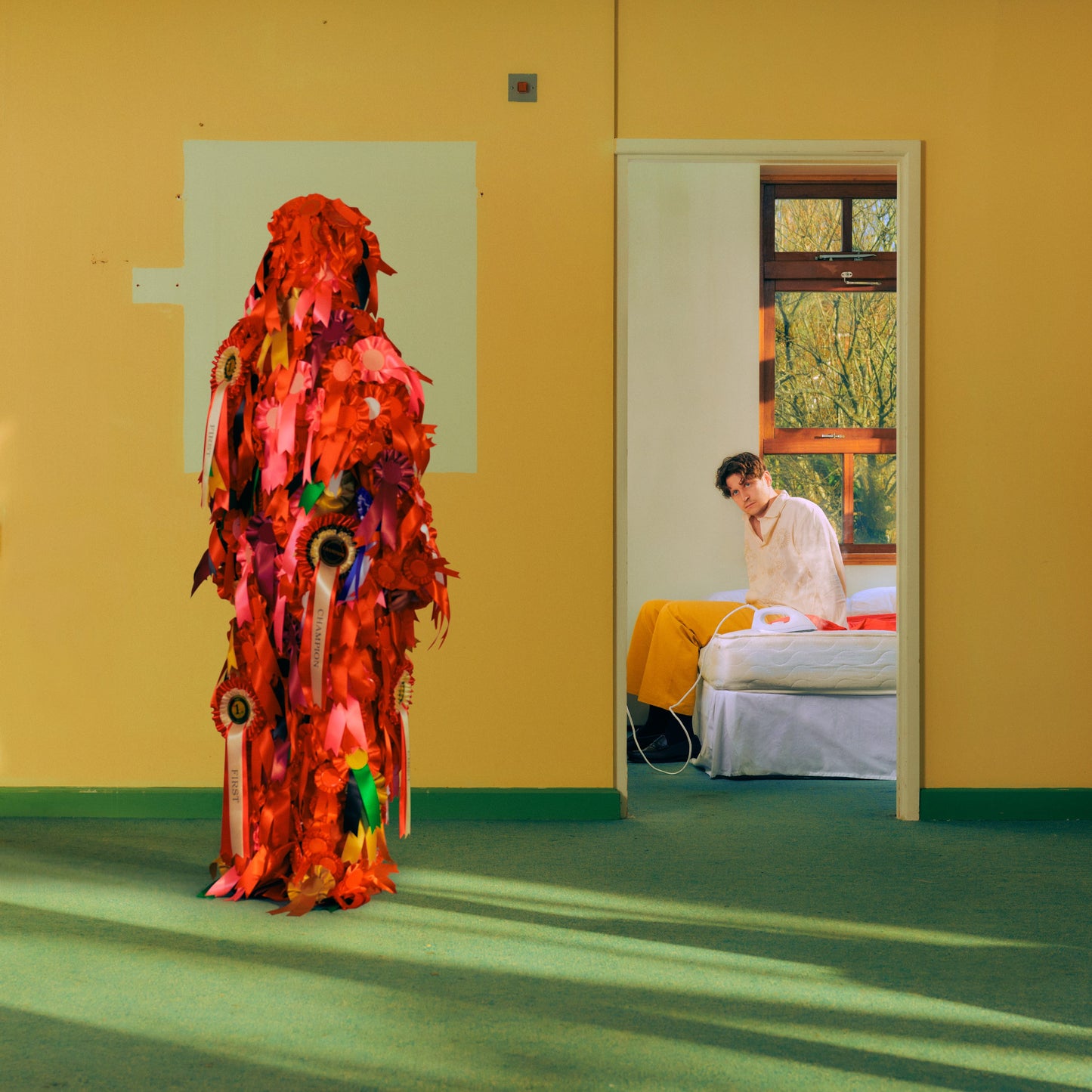 A figure covered in red award ribbons stands before a man sitting on a bed.