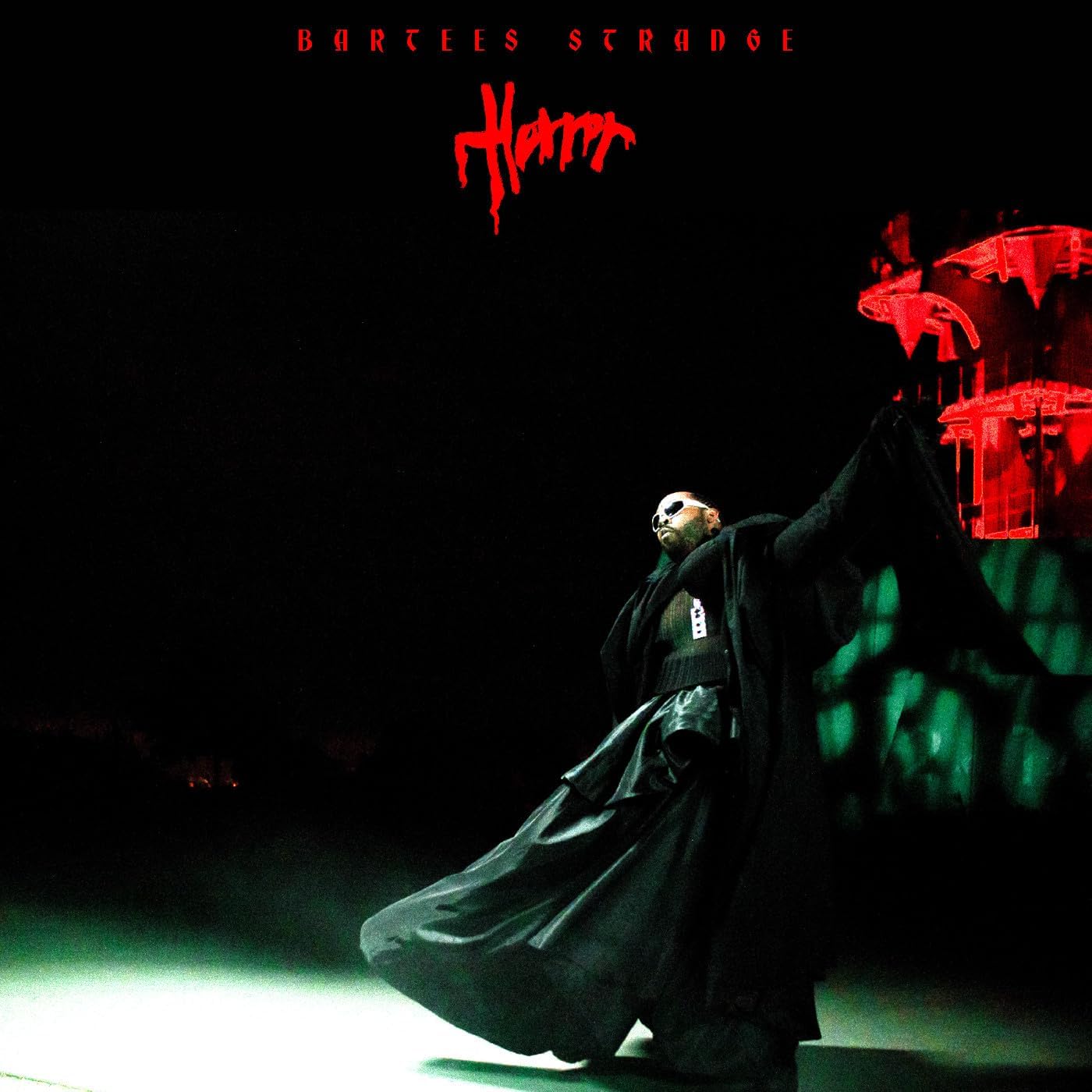 Bartees Strange in a black coat and skirt against a dark, red-lit background.