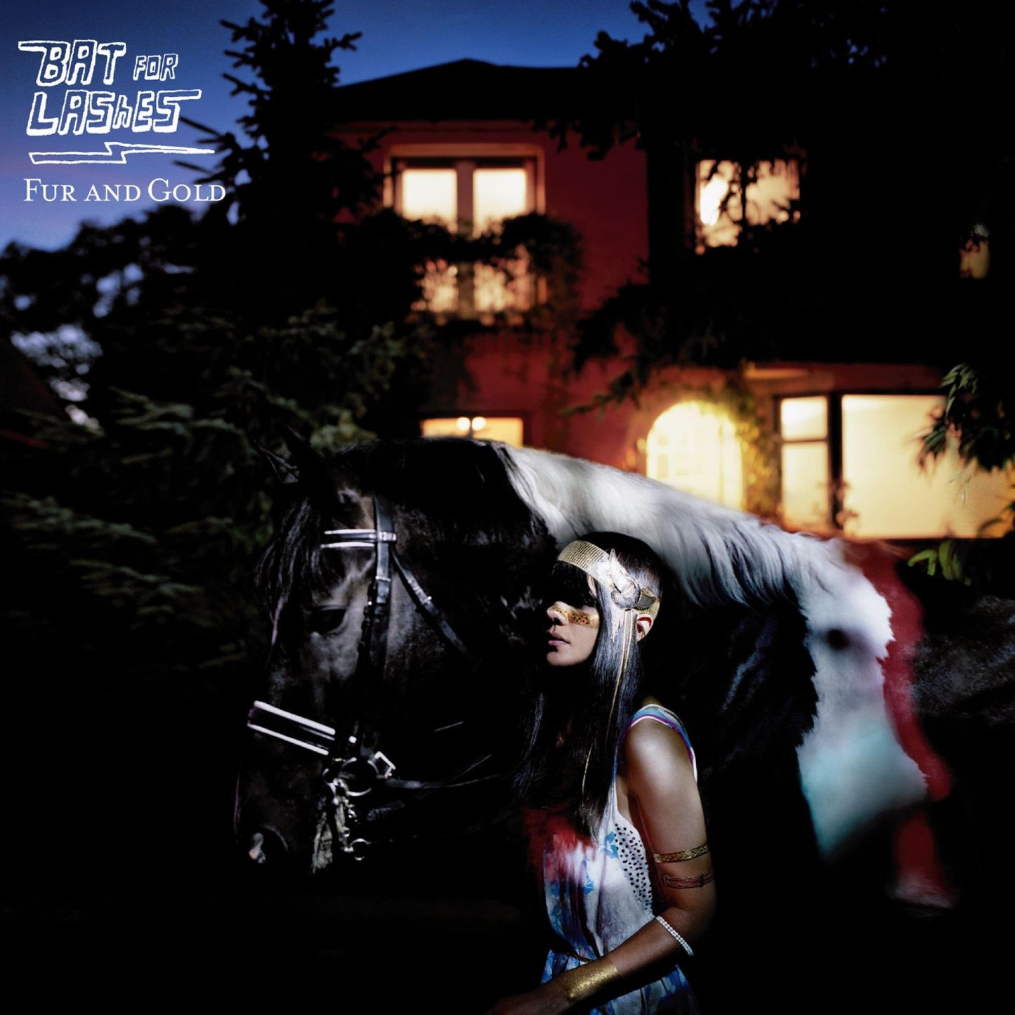 A woman wearing gold headgear stands beside a black and white horse at night.