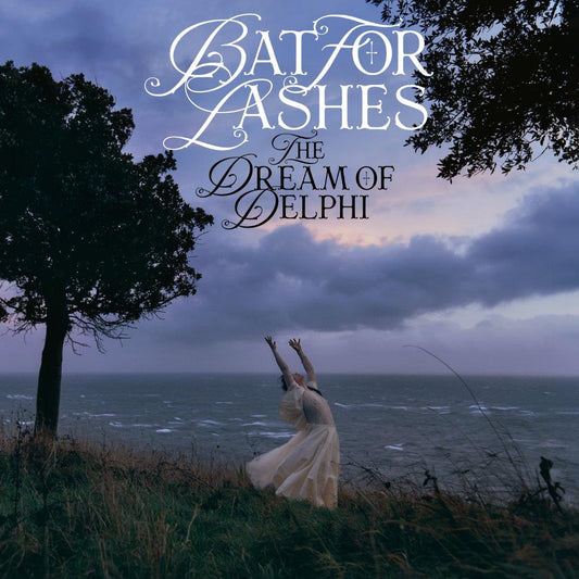 A person in a white dress reaches toward the sky by a stormy ocean.