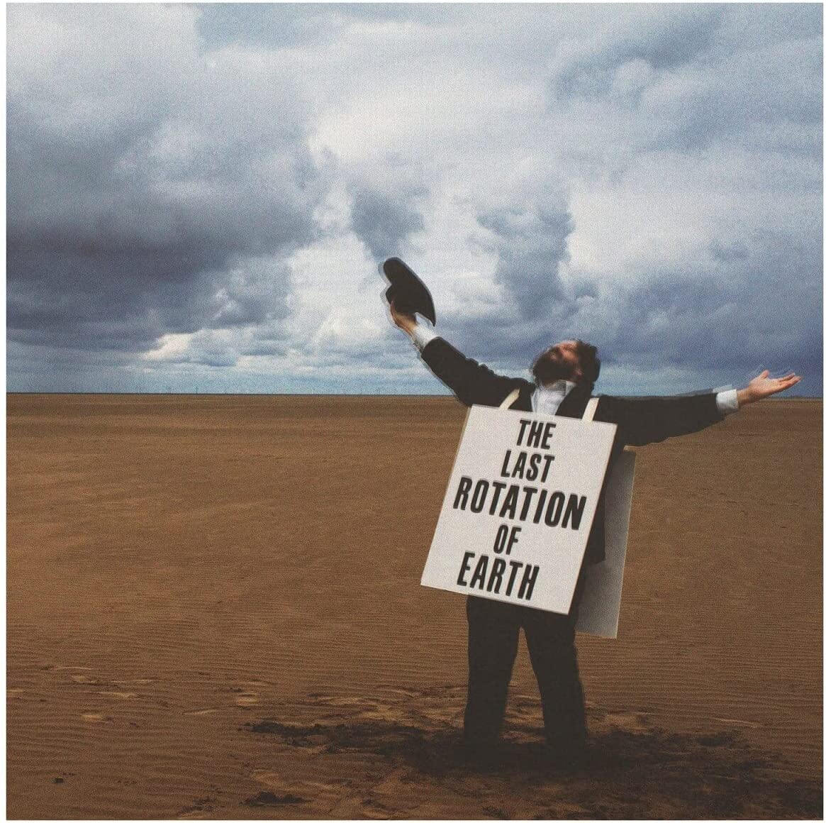 Man in suit stands in desert, holding shoe, with sign reading "The Last Rotation of Earth".