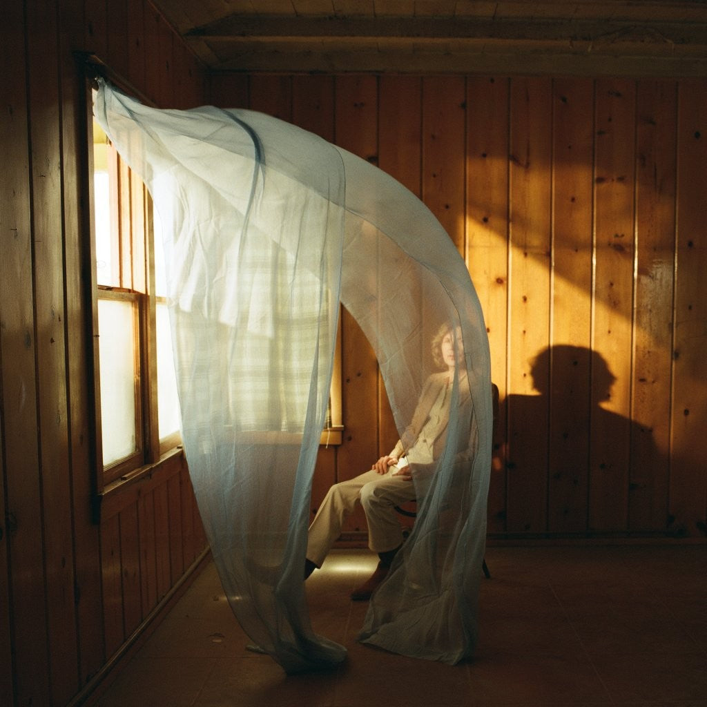 Person seated near a window, draped with sheer fabric, in a wooden room.